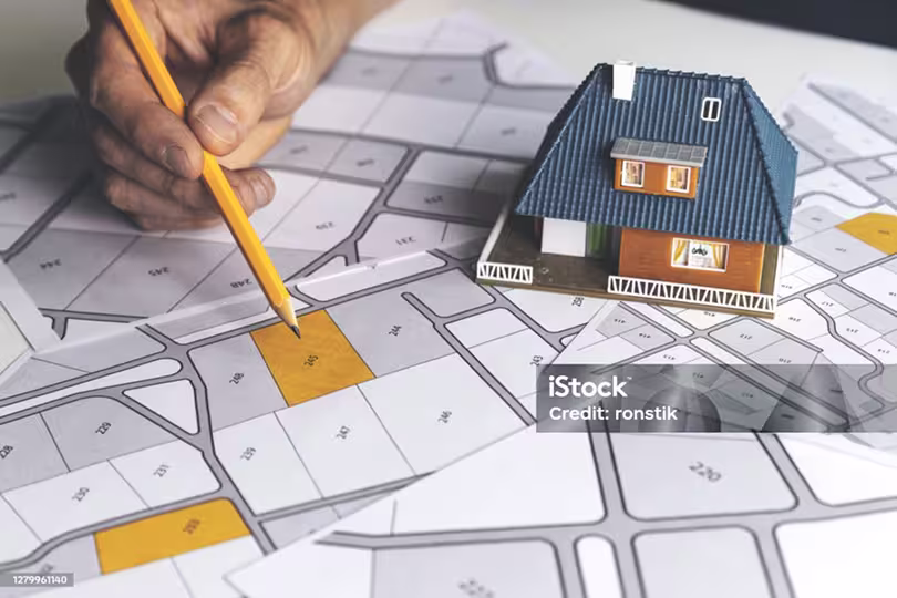 Maketa kuće na detaljnom geodetskom nacrtu dok ruka olovkom precizno označava specifičnu česticu na planu.